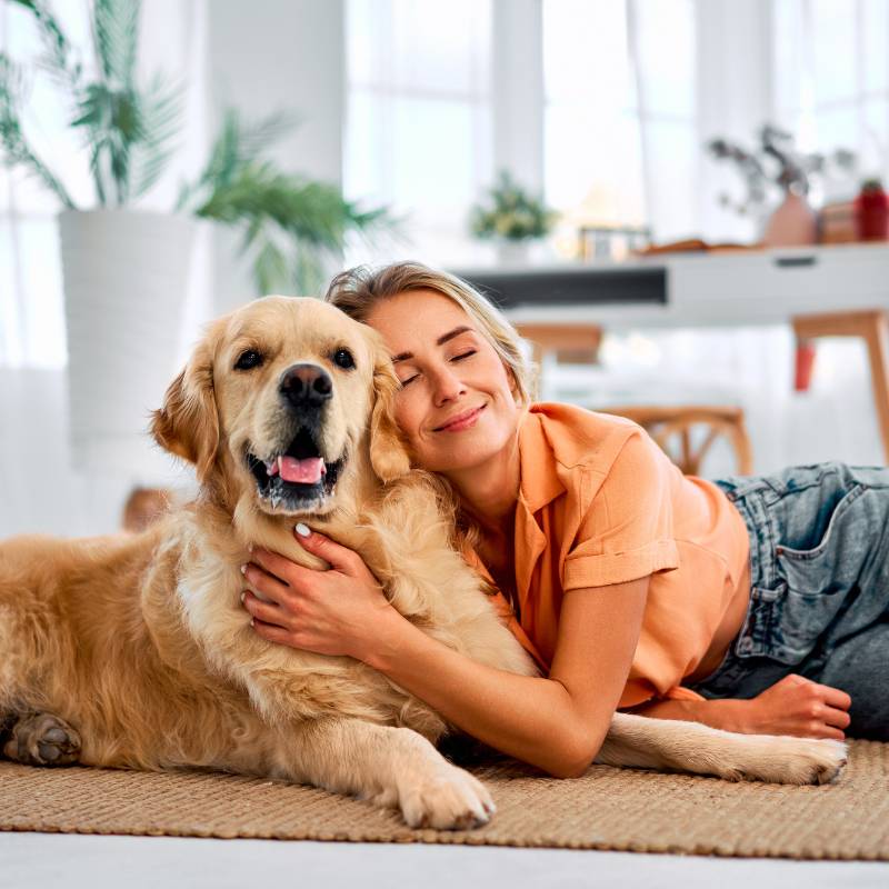 A woman in an orange shirt hugs a golden retriever on a woven rug in a bright room A woman in an orange shirt hugs a golden retriever on a woven rug in a bright room